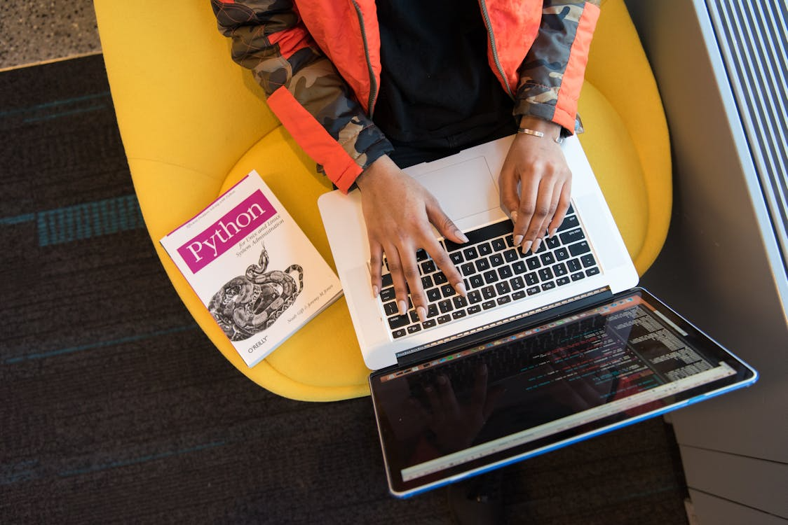 Person typing on a laptop with a Python programming book beside them